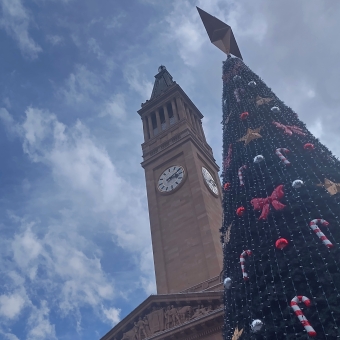 Festive City Hall, Brisbane City, with the City Hall clock tower and a ornate Christmas tree standing against a blue sky which has white clouds occasionally dotted amongst it