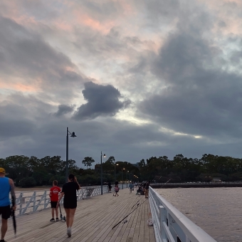Evening Pier, Shorncliffe, with a long and wooden pier with a few people walking along it as well as sone fishing lines off the side into the water beneath it, with grey clouds mixing with pastel pink ones behind them