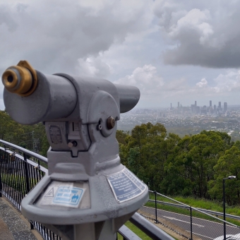 A Closer View, Mt Coot-Tha, with a set of fixed binoculars pointing towards the grey silhouette of Brisbane Central Business District and surrounds, with thick grey clouds above
