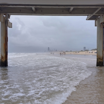 The Spit's Distance, Main Beach, Gold Coast, with the pictre framed with a bridged pipeline going above and its supports left and right, with whitewashed water below, with a long and straight beach disappearing off into the distance and the Gold Coast skyline barely visible beneath thick layers of cloud