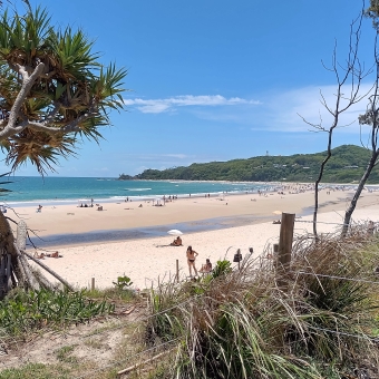 Towards The Lighthouse, Byron Bay, with a tree left and right which frames the image, with a yellow beach in the foreground dotted with many people, with a low-rise hill with a lighthouse dotted atop it, with the clearest blue sky reflected in the water on the left