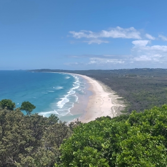 Tallow Beach from Cape Byron, Byron Bay, with bushy trees in the low foreground, with bluest sky above, with water on the left and beach in the centre and dense bush on the right which is viewed from such a height as to give the illusion of carpets sewn together