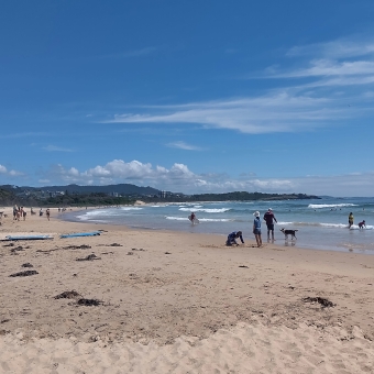 North Wall Beach, Coffs Harbour, with sand on the left and water on the right, with some people sread out across thesand and water, with a low-rise hill covered in trees extending into the water, with bluest sky above