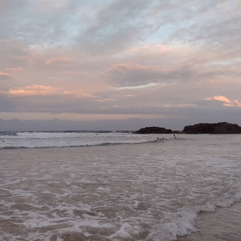 Oxley Beach Sunset, Port Macquarie, with grey clouds above gilded with pastel pink and peach, with whitewashed waves below, with two large boulders outcropping out of the water