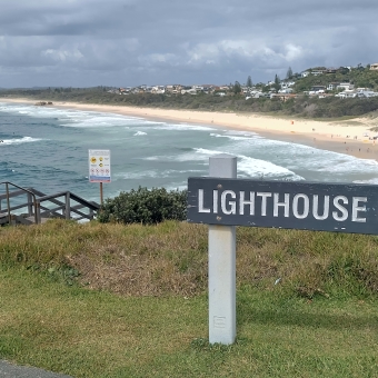 Lighthouse Beach, Port Macquarie, with a wooden sigh in front which says 'Lighthouse Beach' atop a grassy ledge, with blue rolling waves below the hill, with a yellow beach extending left and right, with assorted trees and houses on the low hill above it and blue but cloudy skies above