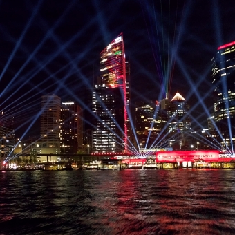 The Well-Lit Nautical Jaunt, Circular Quay, with Circular Quay train station lit up red over the ferry wharfs, with light beams latticed across the sky, with reflections haphazard in the choppy water