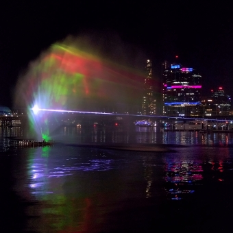 Sprayed Projection, Tumbalong, with a large fan of misted water sprayed out from the water below and lights and lasers projected on it, with the towers of Barangaroo in the far distance