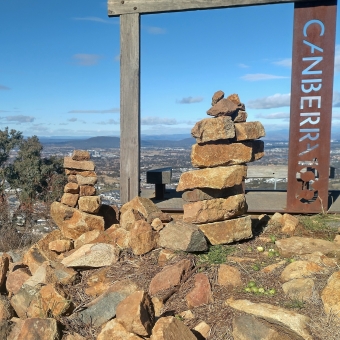 Rock Piles Atop One Tree Hill, Taylor, with three one-metre-tall piles of rocks in the foreground with a frame for Canberra Centenary and one tree in the midground, with a valley with housing development in the background below a clear blue sky
