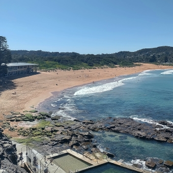 Expanse of Avalon Beach, Avalon, with nothing in the immediate foreground, with rocks half-covered in water then waves over the beach then dunes covered in foliage in front of clear sky