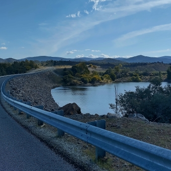 Cotter Saddle Dam, Cotter Dam, with a road along the dam wall in the foreground going to the left and into the distance, with water then low rolling hills in the middle and right of the picture