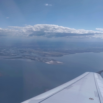 Clouds Above The Coast, from the plane, with a plane wing in the foreground against blue water far beneath leading to flat land and a large gap to a flat layer of cloud