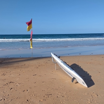 The Flags on Manly Beach, Manly, with a red and yellow lifeguards flag and a kayak with the insignia 'lifeguard' on it and yellow sand leading to low rolling waves coming in and a clear blue ocean and sky