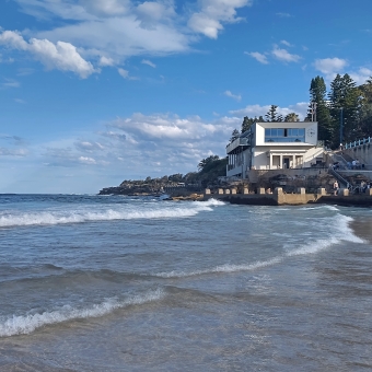 The Pool Club on Coogee Beach, Coogee, with rolling waves left to right and a building overlooking the ocean with a man-made rockpool beneath it, with a blue sky with a small smattering of wispy clouds