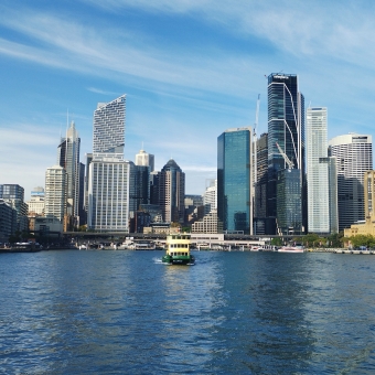 The Bluest View, Circular Quay, Sydney, with a yellow and green ferry swimming towards the camera, with wharves then skyscrapers in front of thin-streaked clouds and blue sky in the background