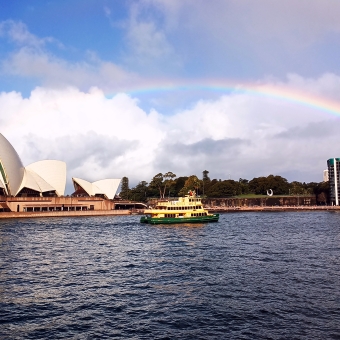 The Rainbow Across Sydney, Sydney Harbour near Bennelong Point, Sydney, with the Sydney Opera House on the left, a yellow and gold Sydney ferry in the centre, and low skyscrapers on the right, with a rainbow arcing in the sky and choppy water below