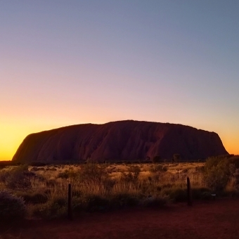 Uluṟu, Sunrise from Sunset Viewing Area, with Uluru emerging from the ground, with yellow and peach and baby-blue glowing from the horizon