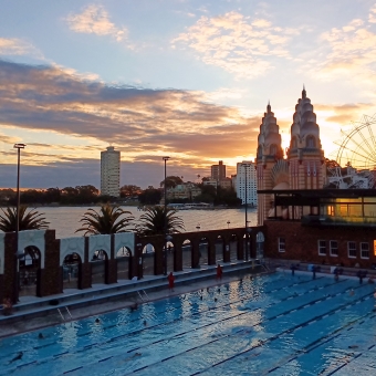 Aquatic Sunset, North Sydney Olympic Pool, Milsons Point, with a glowing sunset amongst streaky clouds hiding behind the Luna Park entrance which itself is behind a pool in shadow underneath a wall separating inside form outside