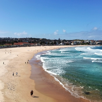 Rolling Waves, Bondi Beach, with sand on the left occasionally dotted with people and waves streaming in from the right, with a low hill dotted with houses and trees in the distance