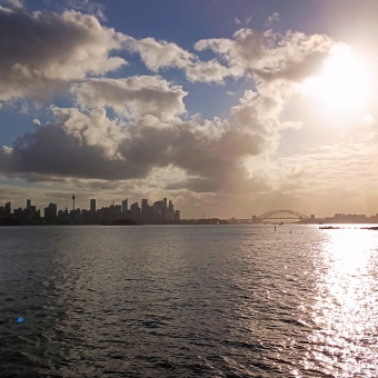 Reflected City, Watsons Bay to Circular Quay Ferry, Sydney, with the Sydney CBD skyline and Harbour bridge silhouetted by a setting sun glowing orange on the right of the picture which contrasts the slightly-cloud-covered blue sky on the left